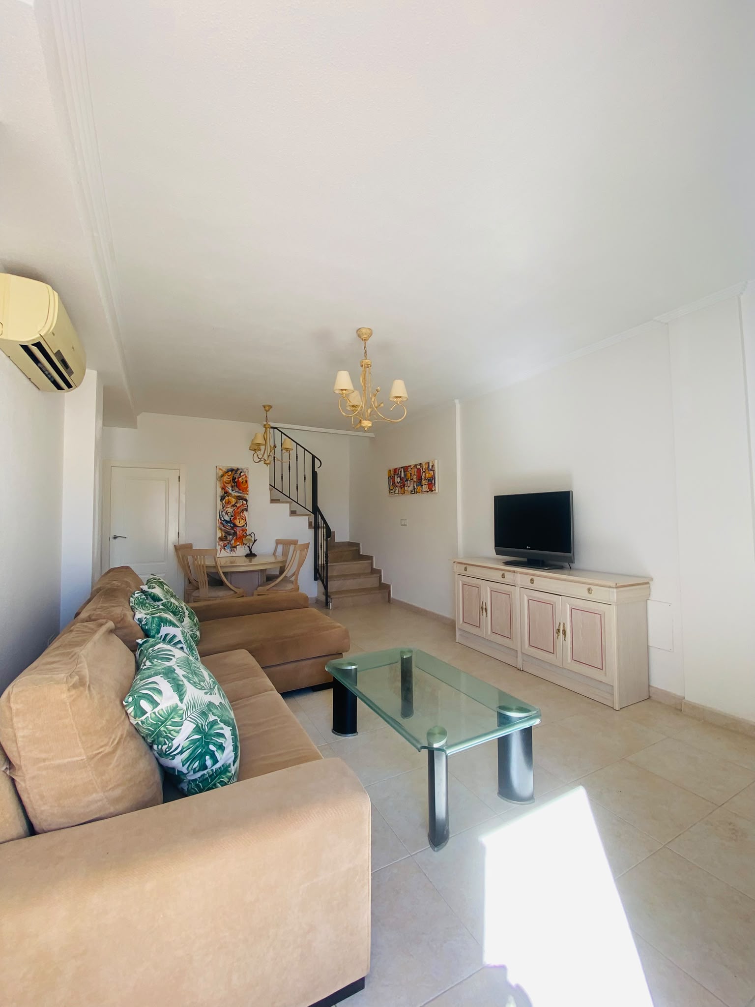 WhatsApp-Image-2026-07-03-at-4.49.39-PM-1 Living room with beige sectional sofa, glass coffee table, TV on a cream cabinet, and a staircase in the background.”,